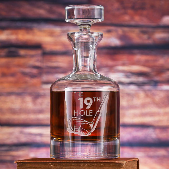 A glass spirits decanter with a faceted stopper, filled with amber spirit, deep-carved with "The 19th Hole" and crossed golf clubs with a ball.