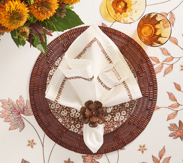 A table setting featuring a floral embroidered tablecloth, brown woven placemat, patterned dinner plate, folded white napkin with brown trim held by a Kim Seybert Begonia Napkin Ring Set of 4, two amber glasses, and a bouquet of orange flowers.