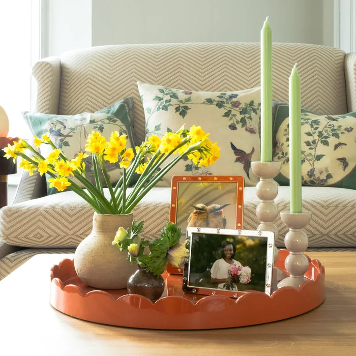 A bright orange tray on a wooden table holds yellow flowers in a vase, two green Addison Ross Bobbin Cappuccino Candlesticks, small framed photos, and a small plant. Floral pillows and a patterned armchair are in the background.