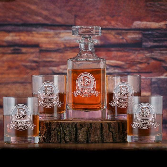 A square whiskey decanter and four rocks glasses, each deep-carved with an initial and name on a circular crest with a banner, filled with bourbon on a wood slab.