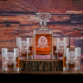 A square whiskey decanter and four rocks glasses, each deep-carved with an initial and name on a circular crest with a banner, filled with bourbon on a wood slab.