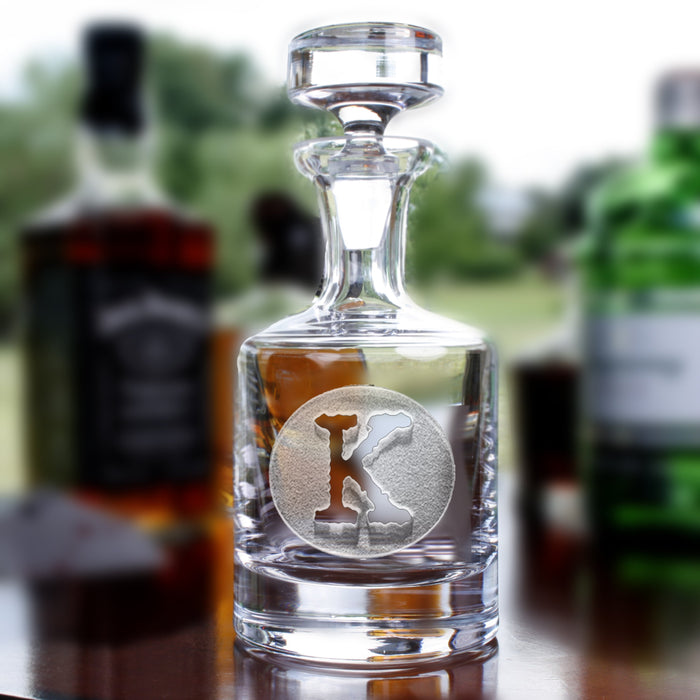 A glass spirits decanter deep-carved with a single block initial "K" in a circle, with whiskey bottles blurred behind.