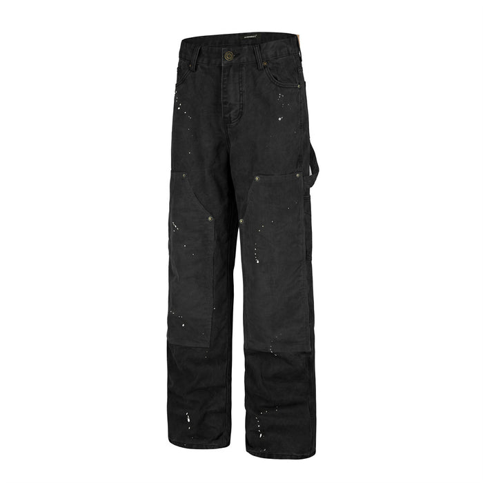 Angled front view of black carpenter pants showing wide legs, cargo pockets, corduroy patches, and white paint splatters