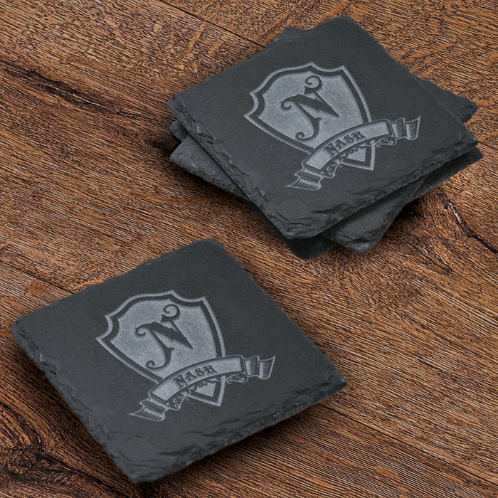 Stack of four square slate coasters deep-carved with a single initial on a heraldic shield above a name banner.
