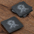 Stack of four square slate coasters deep-carved with a single initial on a heraldic shield above a name banner.