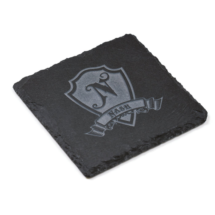 Close-up of one square slate coaster deep-carved with an initial on a heraldic shield and a name banner.