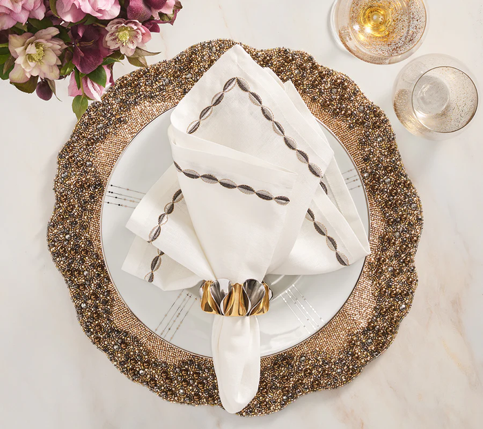 A decorative dinner setting features a gold beaded charger, white plate, embroidered napkin in a Kim Seybert Enigma Napkin Ring (Gold & Silver Set of 4), champagne glass, purple flower bouquet, and elegant gold and silver decor on white marble.