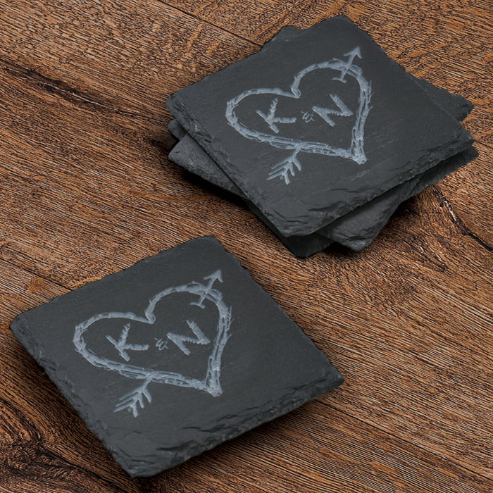 Stack of four square slate coasters deep-carved with two initials inside a rustic heart pierced by an arrow.