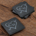 Stack of four square slate coasters deep-carved with two initials inside a rustic heart pierced by an arrow.