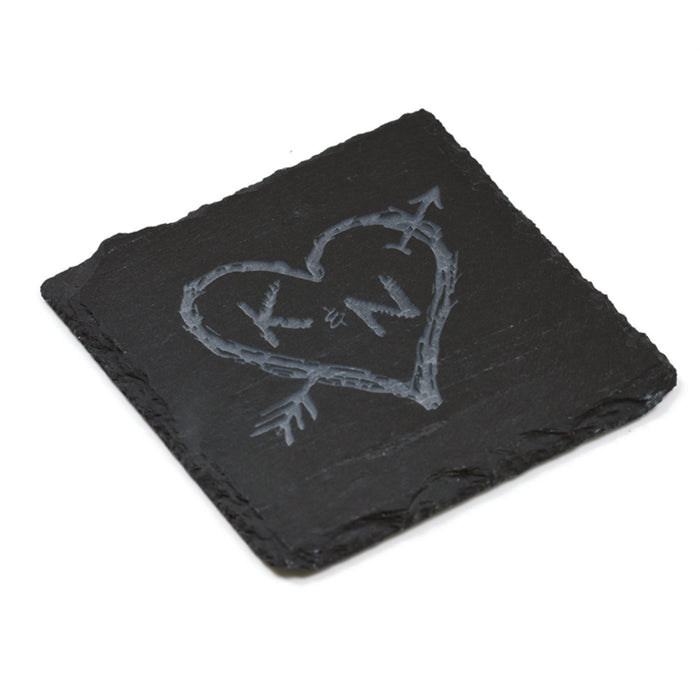Close-up of one square slate coaster deep-carved with two initials in a hand-drawn heart pierced by an arrow.