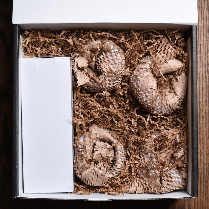 An open white gift box on a dark wood surface, packed with brown crinkle-paper padding, a white inner carton, and bubble-wrapped glassware.