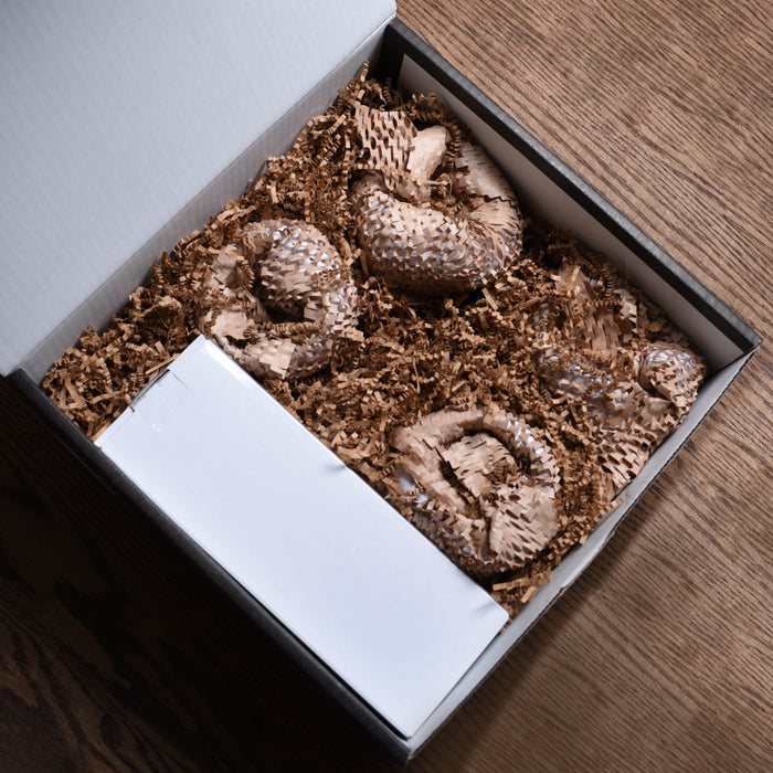 An open gift box seen at an angle on wood, packed with brown crinkle-paper padding, a white inner carton, and bubble-wrapped glassware.