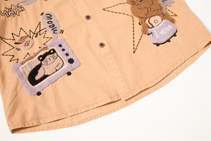 Lower front of the tan shirt showing embroidered cartoon cat graphics, buttons, and finished hem