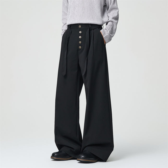 Model wearing black wide-leg pleated trousers, showing the front button fly and long adjustable belt tabs