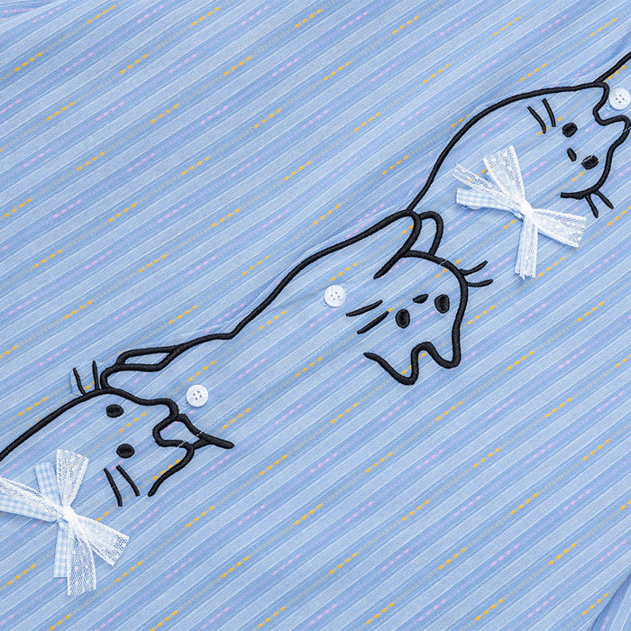 Close-up of three black-outlined cat embroideries with white bows on the light blue striped shirt front