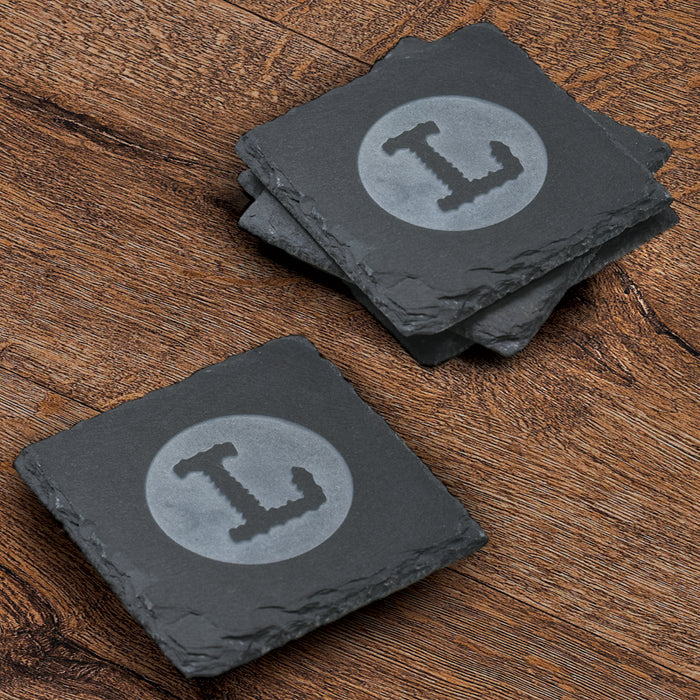 Stack of four square slate coasters deep-carved with a single block-letter initial inside a filled circle.