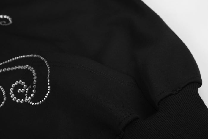 Close-up of a black hoodie sleeve seam with a partial rhinestone scroll design and ribbed cuff