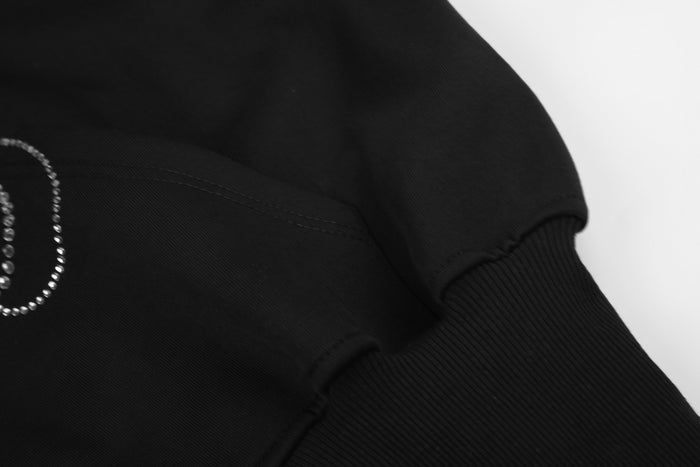 Black hoodie sleeve close-up showing stitched panel seams, a partial rhinestone scroll, and ribbed cuff