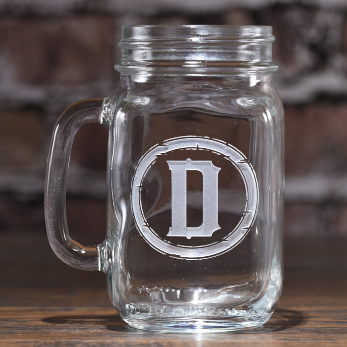 Mason jar mug sand-carved with the initial "D" inside a rustic barbed-wire circle, against a brick wall.