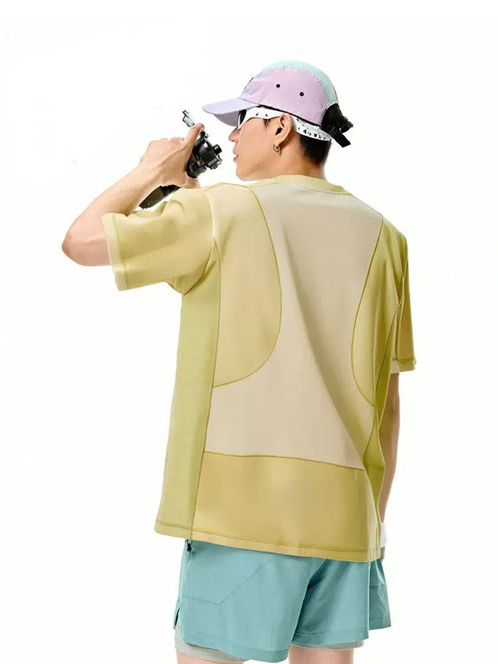 Model wearing a pastel color-block cotton T-shirt with teal shorts and a pink cap, shown from the back holding a camera